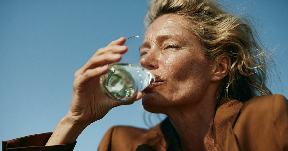 Femme d'une quarantaine d'années buvant un verre d'eau en plein soleil, cheveux au vent