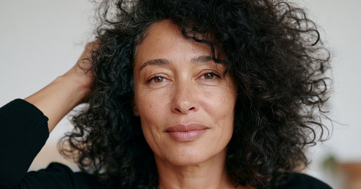 Portrait d’une femme aux cheveux bouclés sombres et fournis, une main dans les cheveux, en lumière naturelle