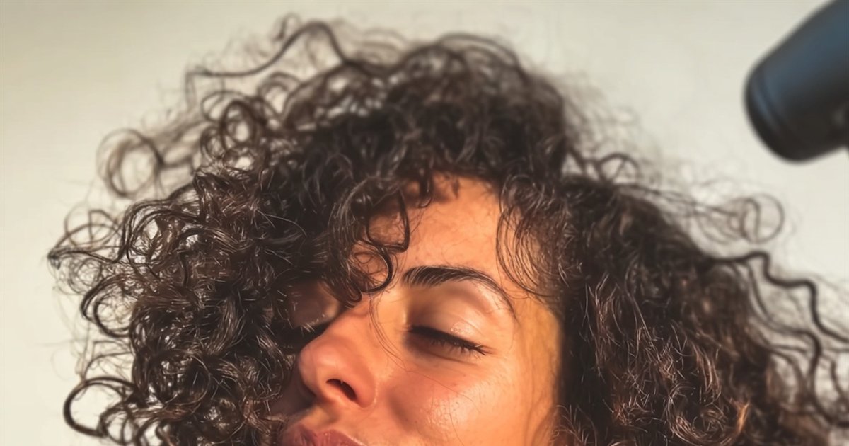Femme brune aux cheveux bouclés, tête légèrement renversée dans la lumière du matin, sèche-cheveux en action