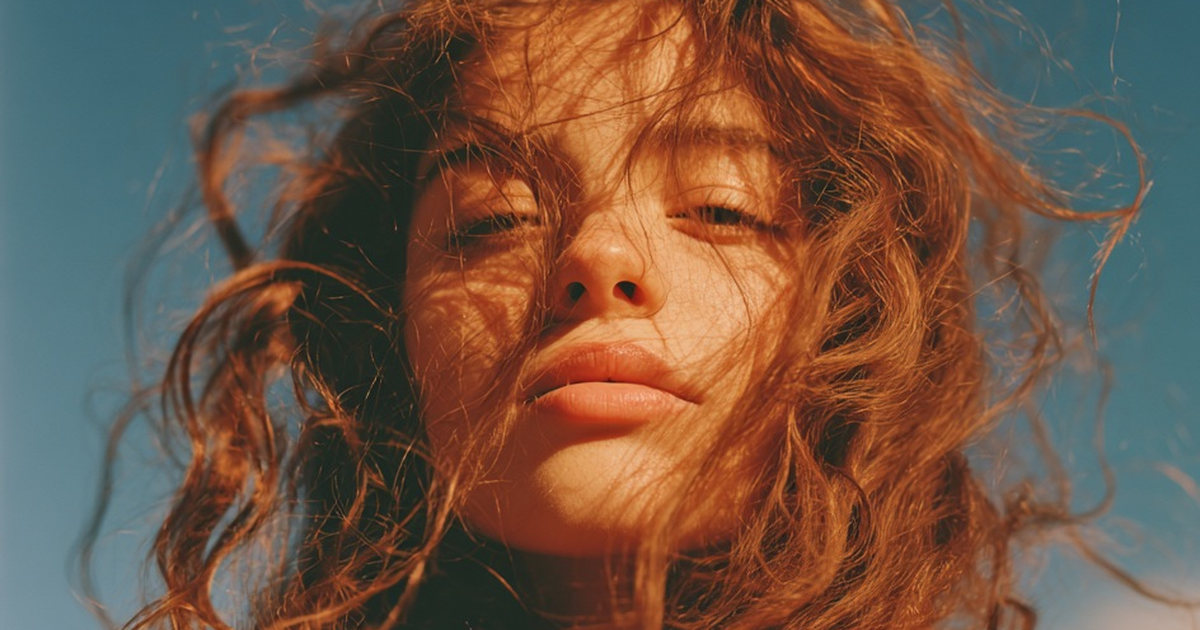 Jeune femme aux longs cheveux bouclés châtain-roux en plein soleil, ciel bleu